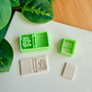 Two light green 3D-printed polymer clay cutters on a white surface. One cutter features an integrated design for a book spine and a whimsical cluster of spotted mushrooms on the cover. Below the tools are two light grey clay samples: one with the embossed mushroom book design and a smaller, plain rectangular piece. Green leaves and a wooden surface are visible in the background.