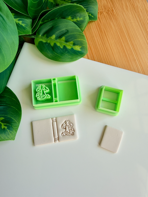 Two light green 3D-printed polymer clay cutters on a white surface. One cutter features an integrated design for a book spine and a whimsical cluster of spotted mushrooms on the cover. Below the tools are two light grey clay samples: one with the embossed mushroom book design and a smaller, plain rectangular piece. Green leaves and a wooden surface are visible in the background.