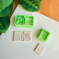 A top-down product flat lay showing a large light green 3D printed polymer clay cutter with a detailed magnifying glass stamp (with 'LM' letters inside the lens) and a book spine design in one chamber, and a simple rectangular cutter in the other. Below it, a rectangular light grey baked clay piece is clearly embossed with the book and magnifying glass design. To the right, a smaller green rectangular cutter and a small, plain rectangular clay piece are visible. Green leaves and a bamboo surface frame the s