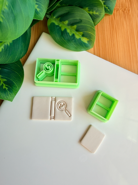 A top-down product flat lay showing a large light green 3D printed polymer clay cutter with a detailed magnifying glass stamp (with 'LM' letters inside the lens) and a book spine design in one chamber, and a simple rectangular cutter in the other. Below it, a rectangular light grey baked clay piece is clearly embossed with the book and magnifying glass design. To the right, a smaller green rectangular cutter and a small, plain rectangular clay piece are visible. Green leaves and a bamboo surface frame the s