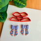 Three light green 3D-printed polymer clay cutters designed for making modular bows, placed on a white surface. Above them are two light grey clay samples: one showing the flat, floral-textured cutout shapes, and another showing a fully assembled 3D clay bow. The background includes green leaves and a wooden bamboo surface.