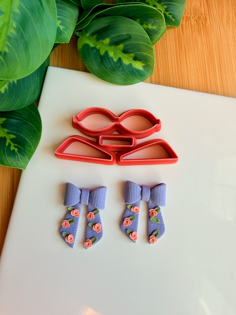 Three light green 3D-printed polymer clay cutters designed for making modular bows, placed on a white surface. Above them are two light grey clay samples: one showing the flat, floral-textured cutout shapes, and another showing a fully assembled 3D clay bow. The background includes green leaves and a wooden bamboo surface.