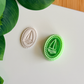 A green 3D-printed sailboat cutter and stamp resting beside a finished white polymer clay charm with a detailed rope border and nautical sailboat design on a minimalist white surface.