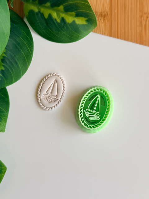 A green 3D-printed sailboat cutter and stamp resting beside a finished white polymer clay charm with a detailed rope border and nautical sailboat design on a minimalist white surface.