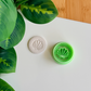 A top-down view of a mint green circular clay cutter with a central scallop shell design and a dotted "pearled" border, sitting next to a white polymer clay medallion that shows the crisp, embossed impression of the same nautical design on a clean white surface with green leaves in the corner.