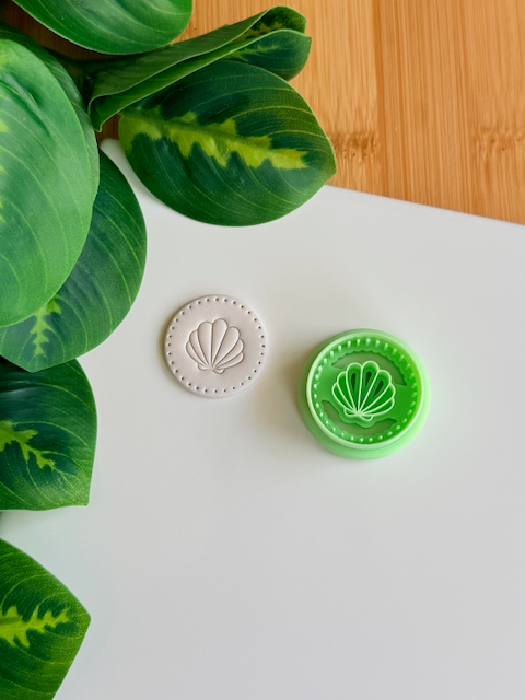 A top-down view of a mint green circular clay cutter with a central scallop shell design and a dotted "pearled" border, sitting next to a white polymer clay medallion that shows the crisp, embossed impression of the same nautical design on a clean white surface with green leaves in the corner.