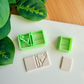 Two light green 3D-printed polymer clay cutters on a white surface. One cutter features an integrated design for a book spine and an elegant "love" script with a small heart on the cover. Below the tools are two light grey clay samples: one featuring the embossed romantic book design and a smaller, plain rectangular piece. Green leaves and a wooden surface are visible in the background.