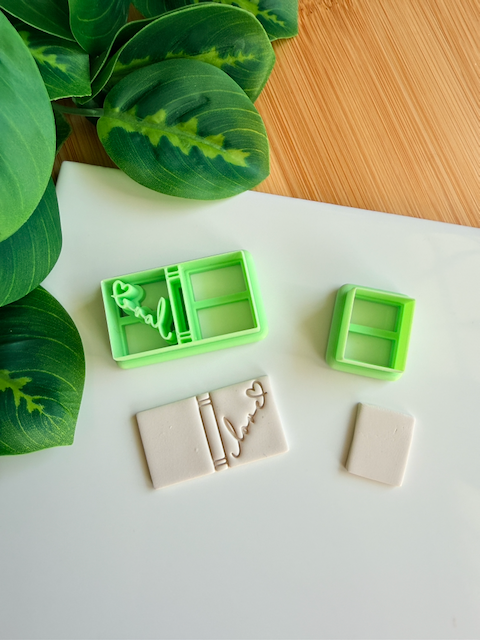 Two light green 3D-printed polymer clay cutters on a white surface. One cutter features an integrated design for a book spine and an elegant "love" script with a small heart on the cover. Below the tools are two light grey clay samples: one featuring the embossed romantic book design and a smaller, plain rectangular piece. Green leaves and a wooden surface are visible in the background.