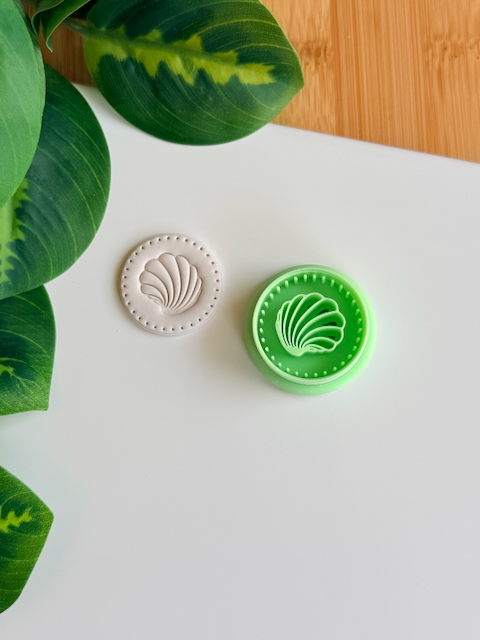 Seashell clay cutter and its white clay impression on a white surface next to green leaves.
